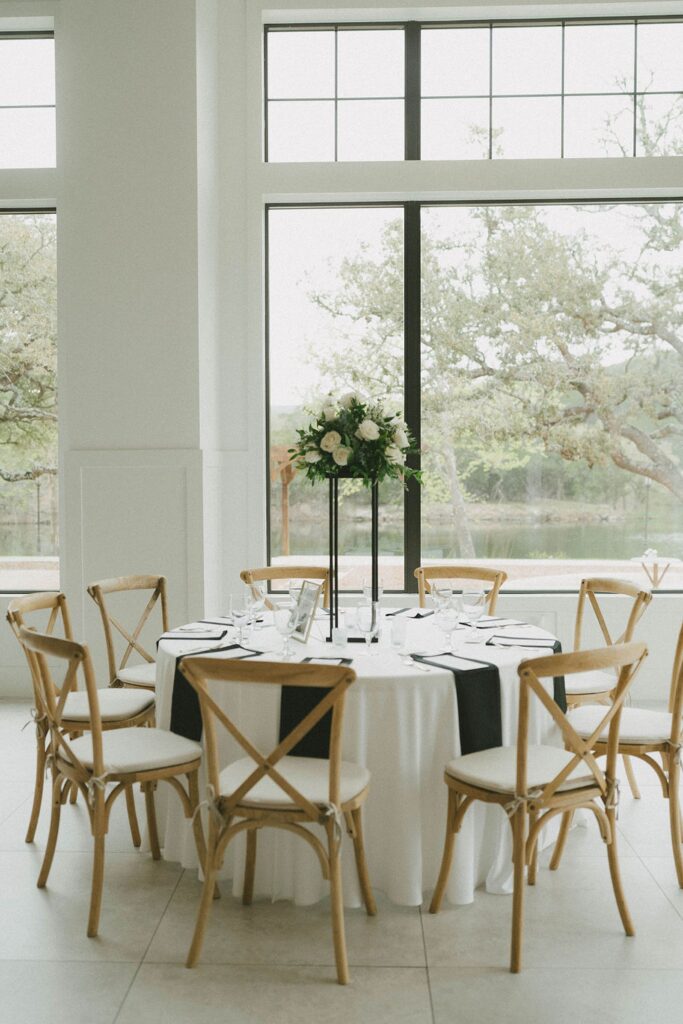 A round table at The Preserve at Canyon Lake is decorated for a formal event with a white tablecloth, black napkins, and a tall floral centerpiece. Six wooden chairs surround the table, while large windows provide a scenic outdoor view.