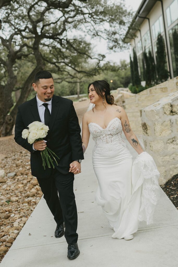 A bride and groom walk hand in hand outdoors on a stone pathway at The Preserve at Canyon Lake, with the groom holding a bouquet of white roses and the bride lifting her dress slightly off the ground.