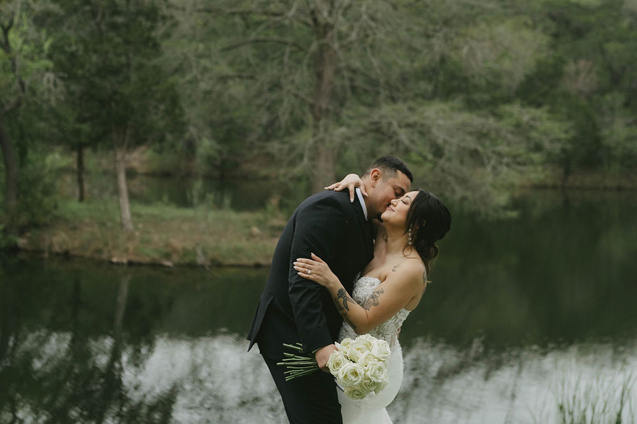 A couple embracing near a serene lake at The Preserve at Canyon Lake, with the woman in a white strapless dress holding a bouquet of white roses and the man in a black suit. Trees are visible in the background.