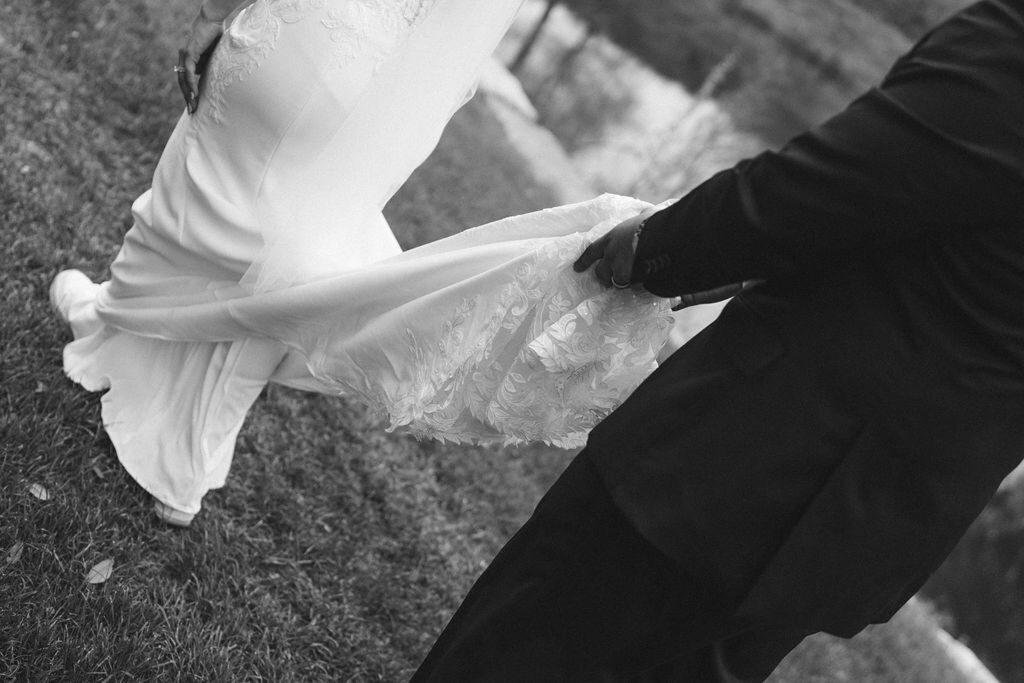 The groom in a dark suit helps lift the bride's white dress as they walk on grass near a water body, focusing on the details of the lace fabric and their lower bodies.