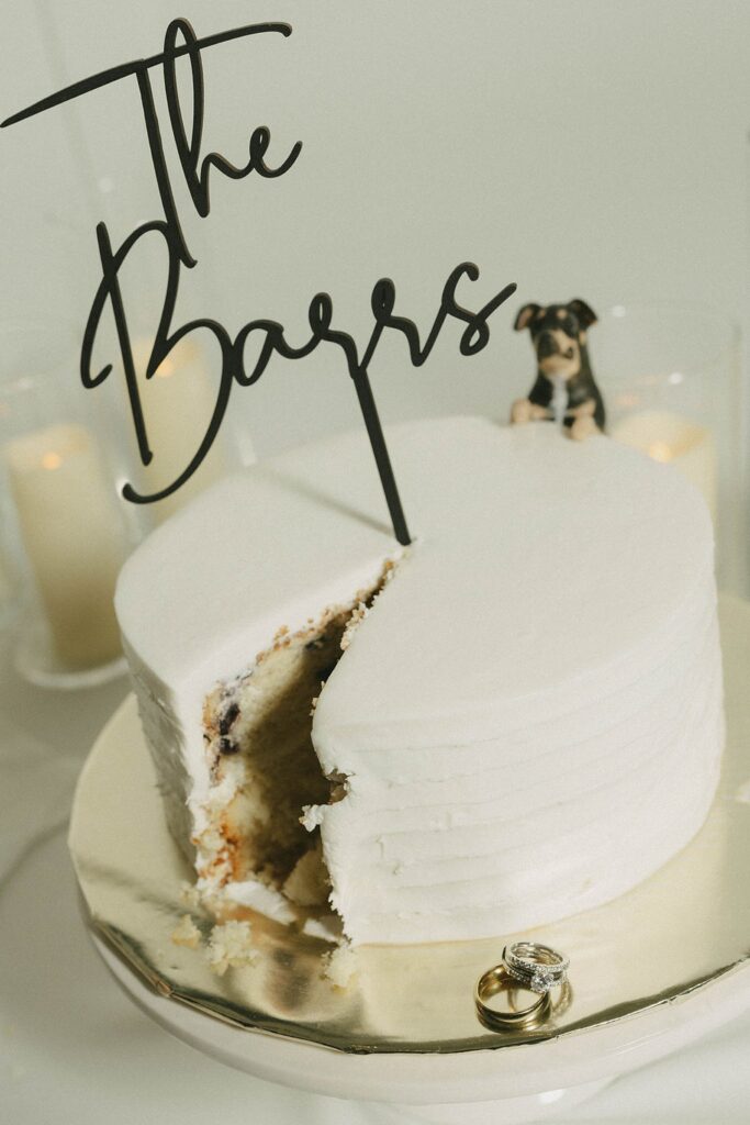 A white frosted cake with a slice removed, a sign reading "The Bagrs" on top, and a dog figurine. Two rings are placed on the cake stand, and candles are in the background.