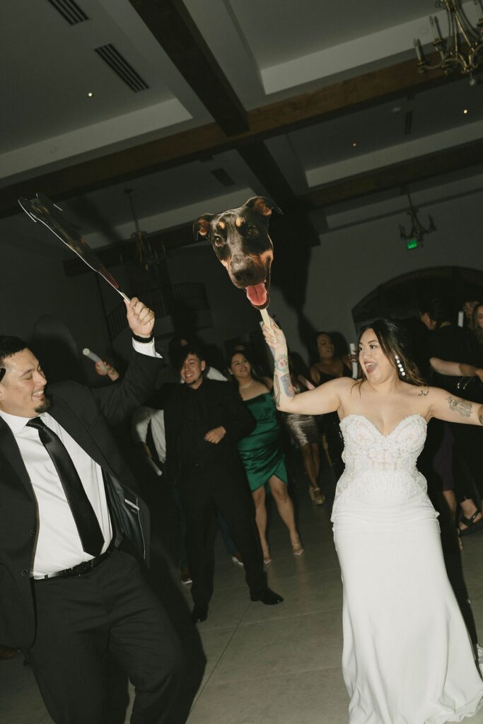 A bride and groom dance together at their wedding reception, each holding a prop with a large cutout of a dog’s face on sticks. Guests are laughing and dancing in the background.