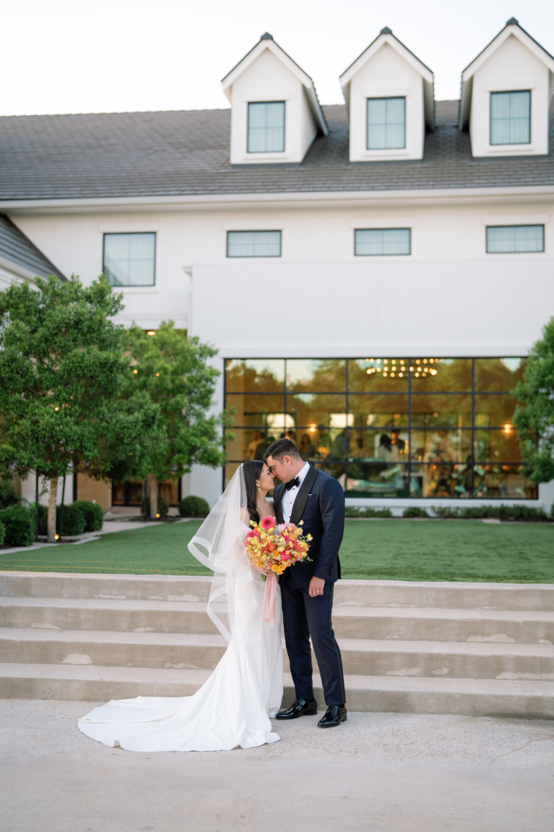 A bride and groom stand on outdoor steps, embracing and holding a bouquet, in front of a modern building with large windows and greenery—capturing the elegance often found at Outdoor Wedding Venues in Texas.