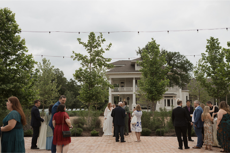 A group of people in formal attire converse outdoors near a large white house, with string lights overhead and trees surrounding the area—capturing the charming ambiance often found at Outdoor Wedding Venues in Texas.