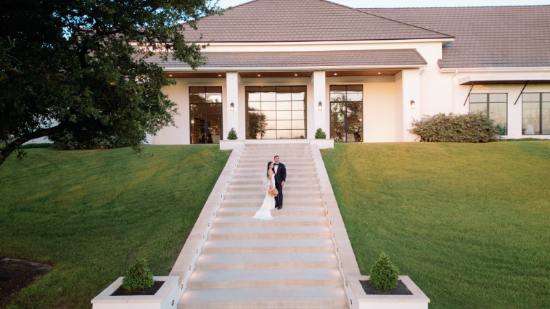 A bride and groom stand together on wide steps leading to a large modern building with tall windows, surrounded by green grass and trees—an elegant example of the scenic Outdoor Wedding Venues in Texas.