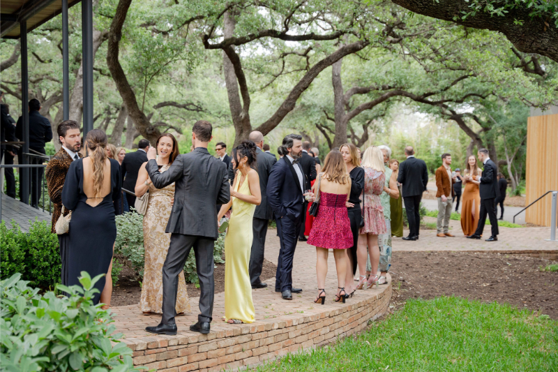 A group of people dressed in formal attire stand and converse outside on a brick path surrounded by trees and greenery, capturing the elegant ambiance often found at Outdoor Wedding Venues in Texas.
