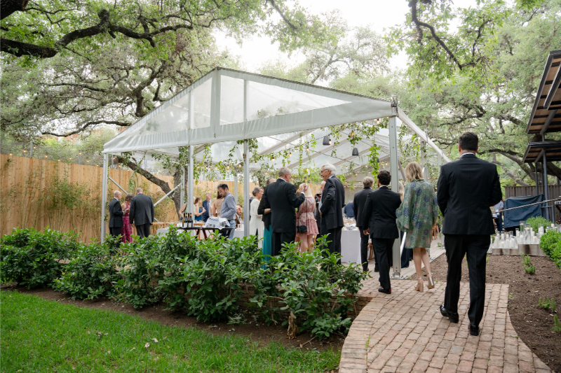 Guests gather under a clear tent for an outdoor event at one of the most charming Outdoor Wedding Venues in Texas, surrounded by lush greenery and brick walkways, with people dressed in formal attire.
