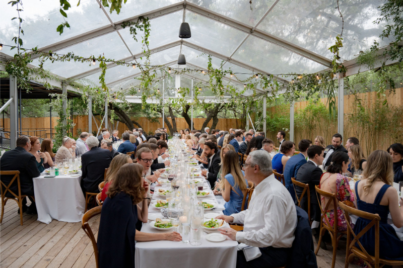 A large group of people sit at long tables under a clear tent, eating dinner at an outdoor event with greenery decorations—capturing the charm often found at Outdoor Wedding Venues in Texas.