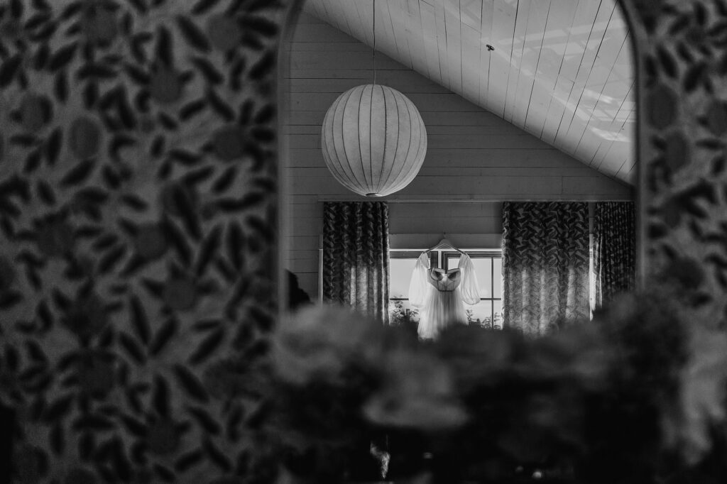 Black and white image of a white dress hanging in front of a window, reflected in a mirror. The room, likely prepared by an Austin Wedding Coordinator, has patterned curtains, a spherical hanging light, and a sloped ceiling.