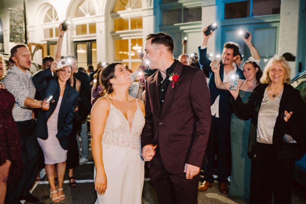 A newlywed couple stands smiling at each other surrounded by people holding phones with flashlights in front of a building at night.