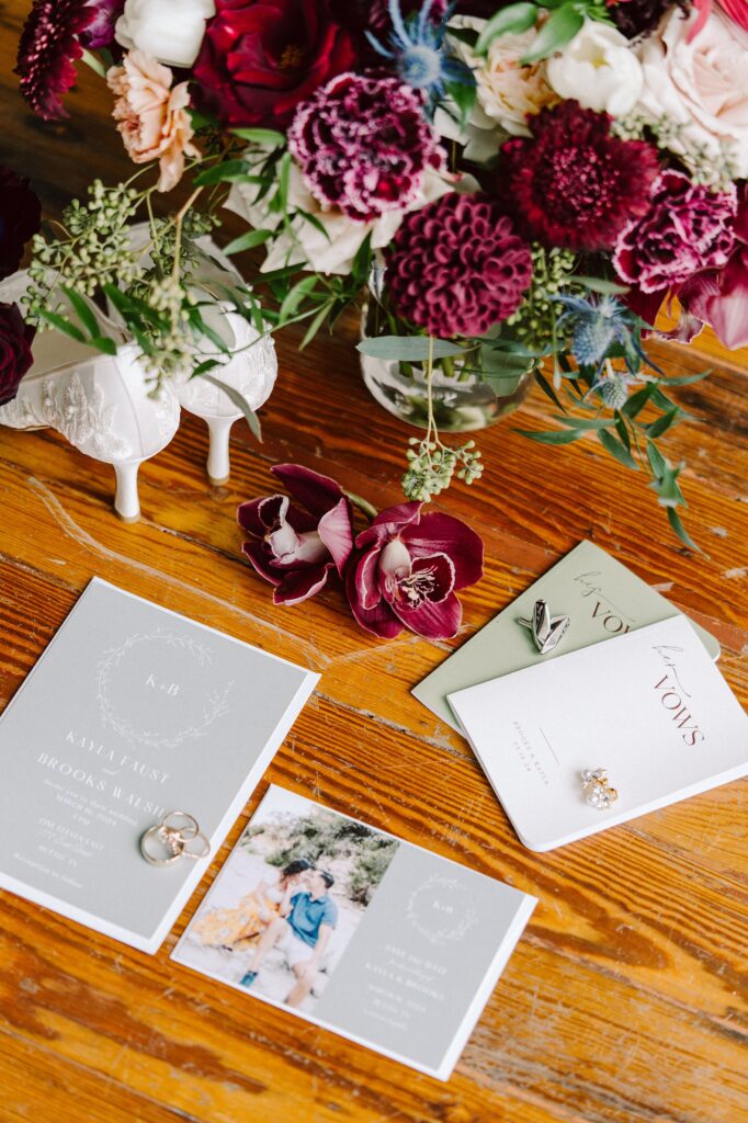 Floral arrangement, wedding invitations, rings, and shoes are displayed on a wooden surface. A photo and vow books are also seen.