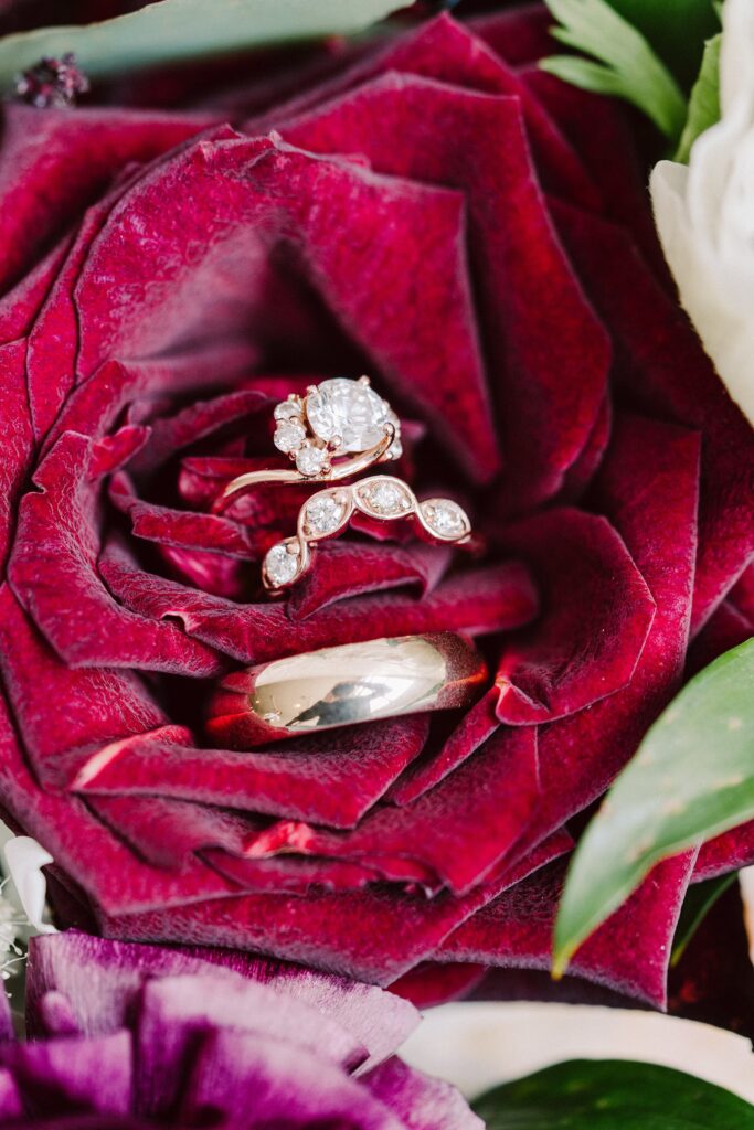 A set of wedding rings, including an engagement ring with a prominent diamond, are displayed on a deep red flower.