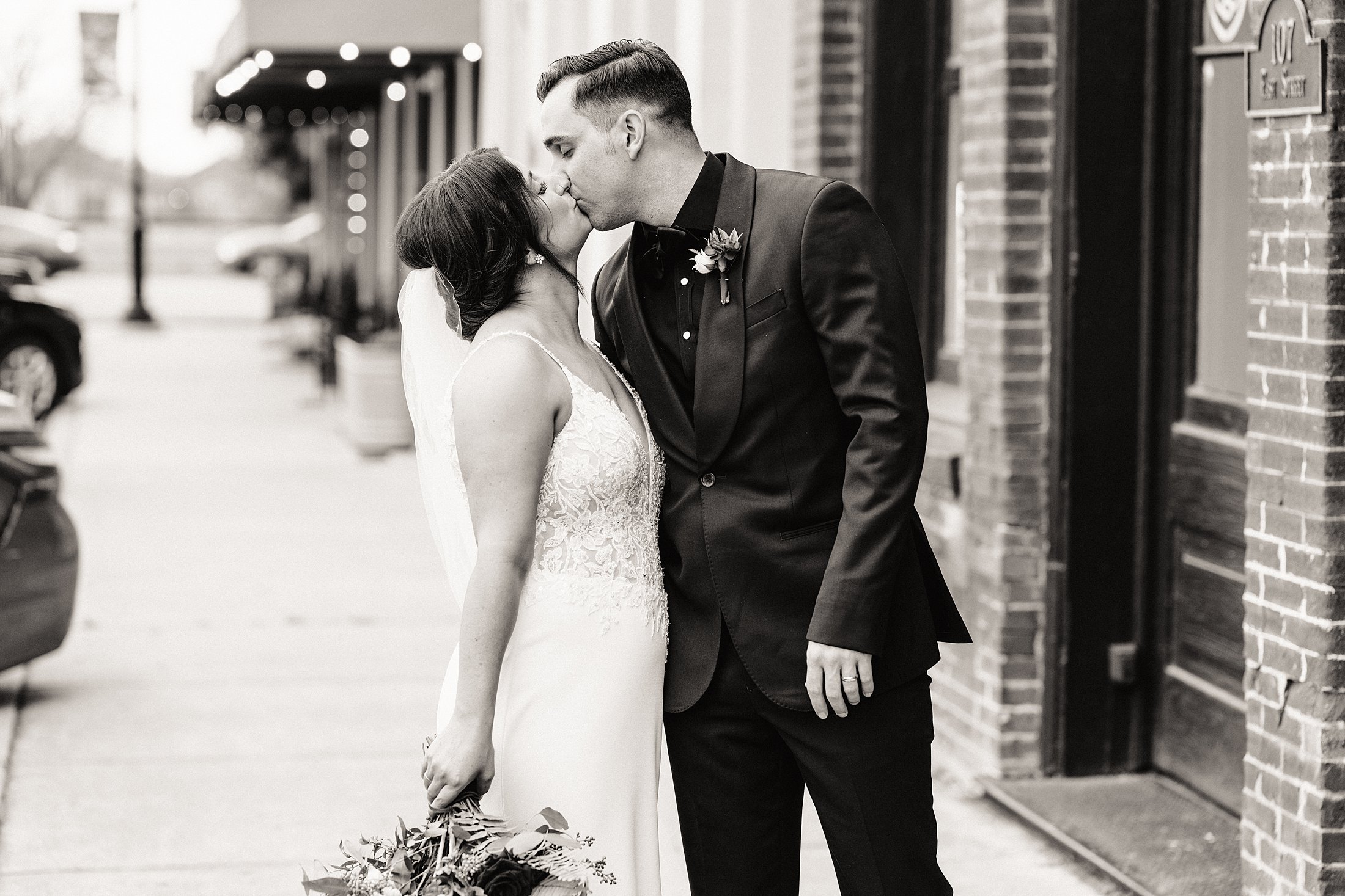 A couple dressed in wedding attire shares a kiss on a city sidewalk. The bride holds a bouquet and wears a veil, while the groom is in a dark suit. The setting appears to be outside of a building.