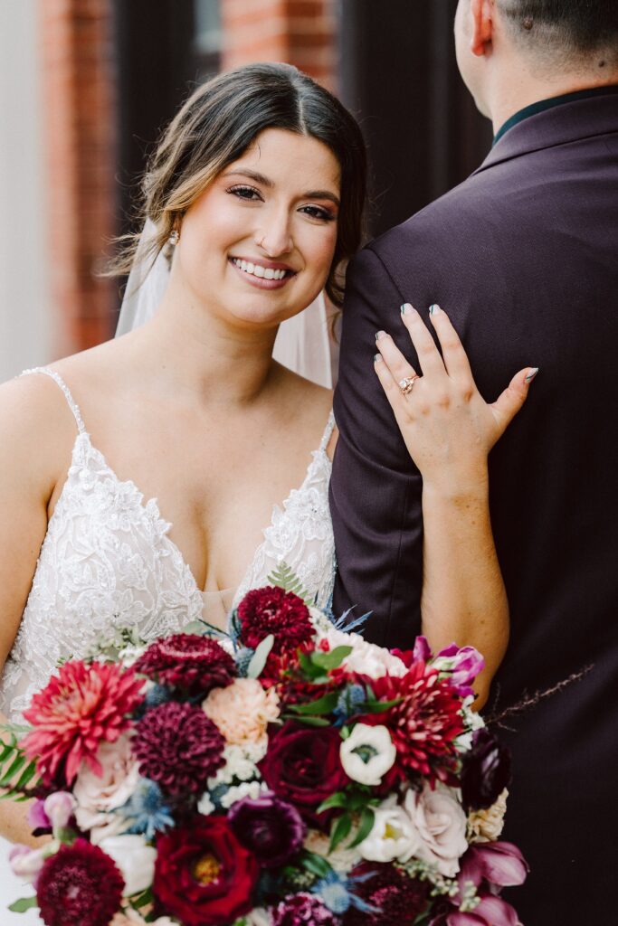 A bride in a white gown holds a bouquet of colorful flowers and places her hand with a wedding ring on the arm of a groom in a dark suit.