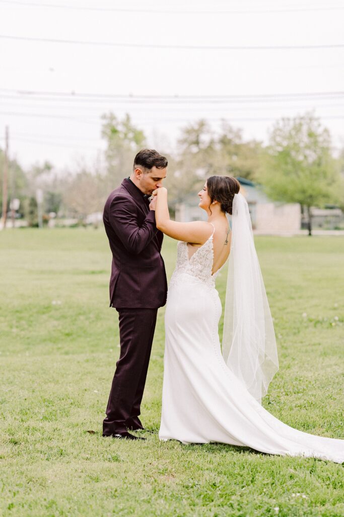 A couple dressed in wedding attire stands on a grassy field. The groom kisses the bride's hand while she smiles at him. Trees and power lines are visible in the background.