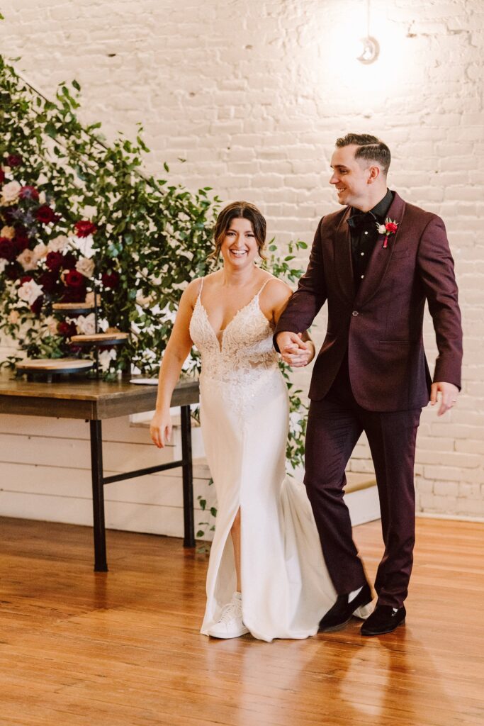 A bride in a white dress and a groom in a burgundy suit walk hand-in-hand, smiling, in a venue with a brick wall and floral decorations.