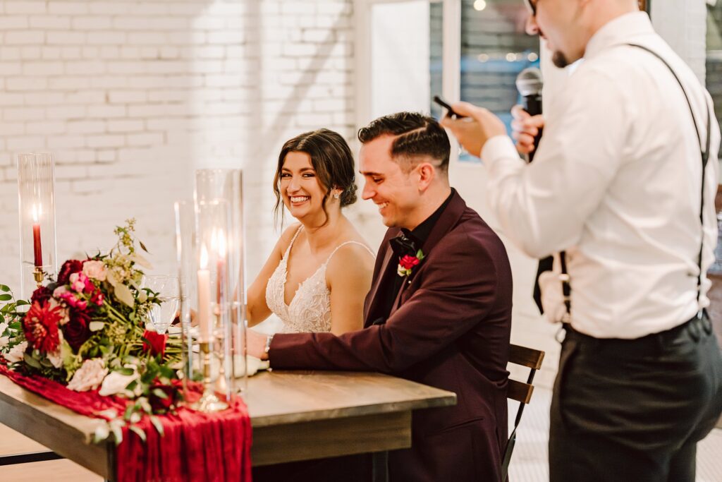 A smiling couple sits at a decorated table during a wedding reception while a man in a white shirt and black suspenders speaks into a microphone beside them.