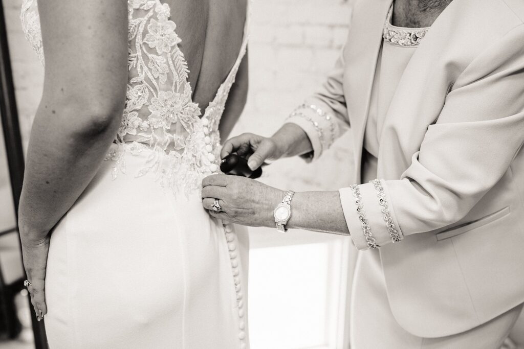 A close-up in black and white of a person buttoning the back of a bride's intricate lace wedding dress.