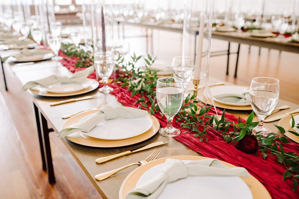 A banquet table is set with white plates, folded napkins, gold cutlery, and glasses. A red runner with green foliage and red flowers decorates the center. Bright, natural light fills the room.