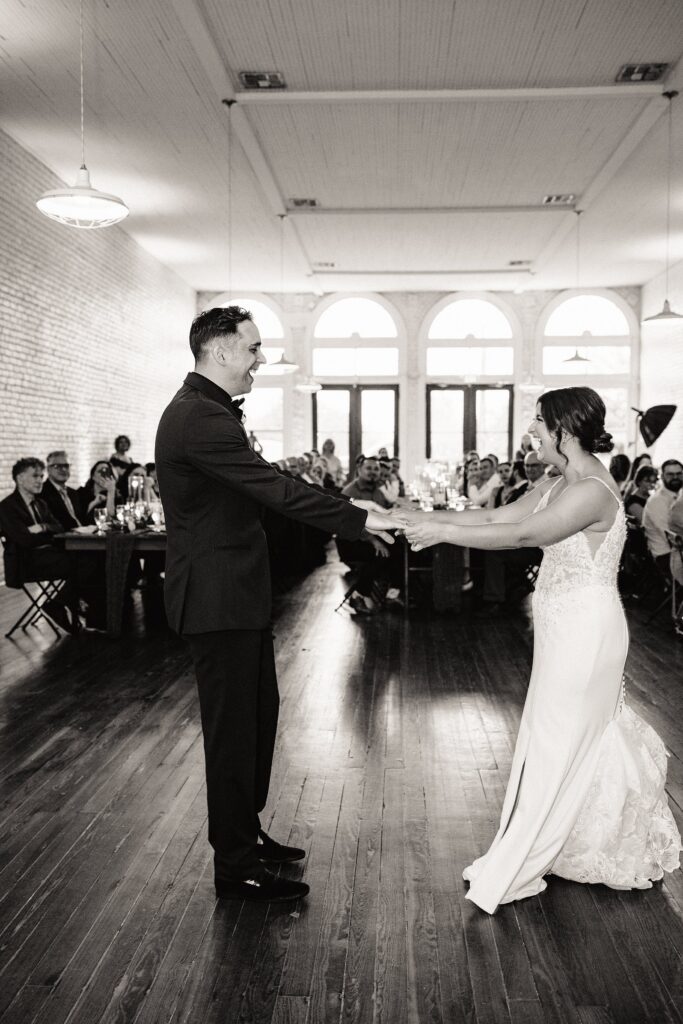 A couple in formal attire dances in a large, bright room with wooden floors and arched windows. Seated guests watch and smile in the background.