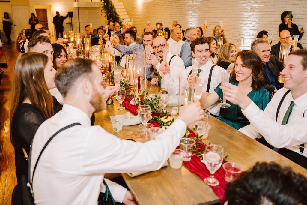 A group of people seated at a long wooden table raises glasses in a toast at a festive event in a warmly lit room, decorated with candles and greenery.