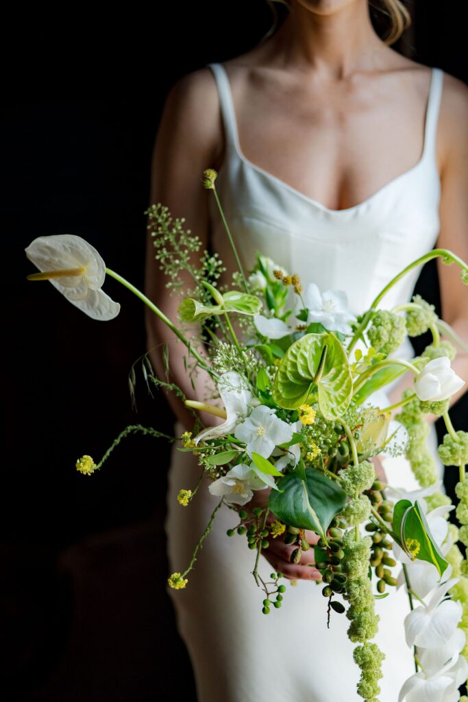 A person in a white dress holds a large, elaborate bouquet of green and white flowers against a dark background.