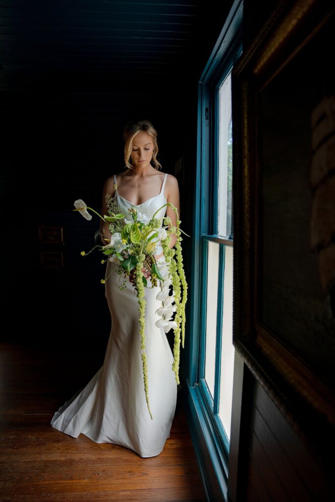 A woman in a white dress stands by a window holding a cascading bouquet of green and white flowers. She looks down, illuminated by natural light from the window. The room is dark.