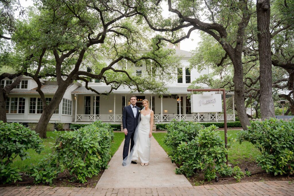 A bride and groom walk hand in hand down a path in front of a large, white, two-story house surrounded by trees and greenery at Mattie's Austin.