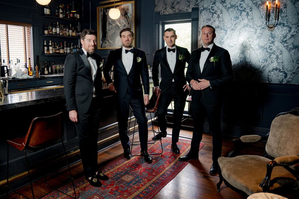 Four men in black tuxedos stand in an elegant room with dark walls, a bar with bottles, and a patterned rug.