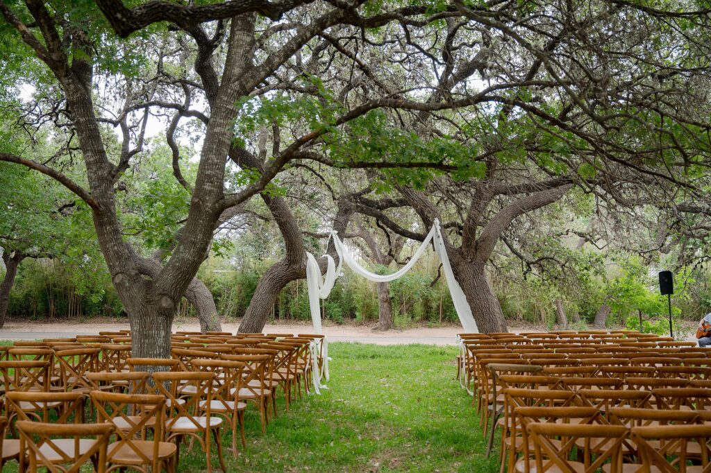 Outdoor wedding ceremony setup with wooden chairs arranged in rows facing an arch draped with white fabric, surrounded by large trees.