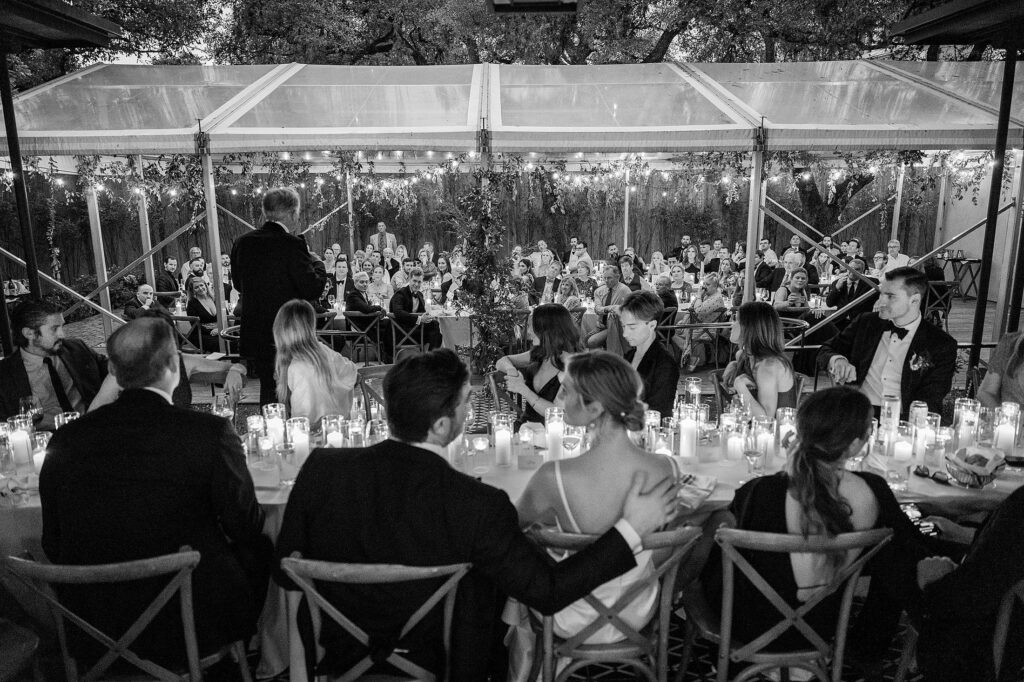 A person gives a speech at a wedding reception in a tent with candle-lit tables and seated guests attentively listening.