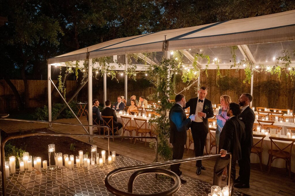 People are gathered under a canopy adorned with greenery and lit by numerous candles, some seated at tables and others standing and talking, in an outdoor evening event setting at Mattie's Austin.