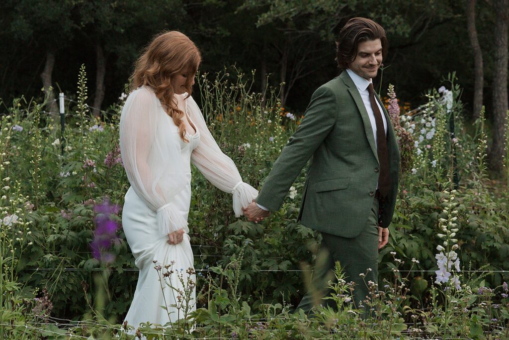 A couple, with one wearing a white dress and the other in a green suit, holds hands while walking through a garden with tall greenery and flowers, beautifully organized by an Austin Wedding Planner.