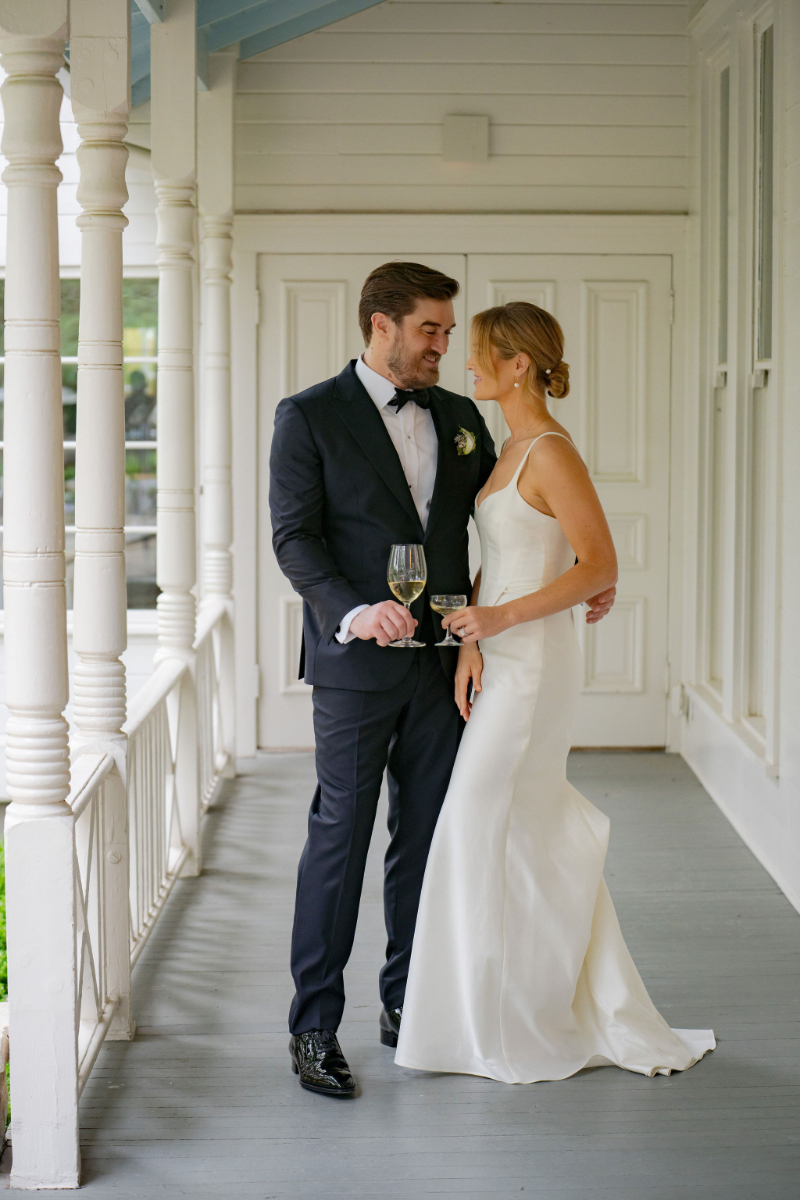 A bride in a white gown and a groom in a dark suit stand on the porch at Mattie's Austin, holding wine glasses and looking at each other.