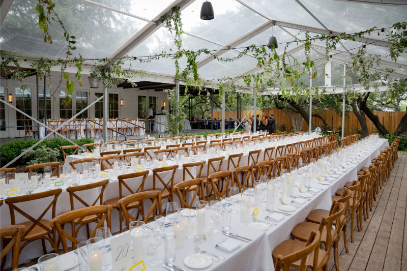 Long tables with white tablecloths, wooden chairs, and glassware are set for an event at Mattie's Austin beneath a clear tent adorned with lush greenery.