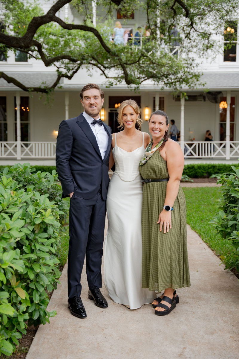 Syd from In Ink Weddings, and Austin wedding planner and design studio, stands next to bride and groom on a garden path in front of a white house at Mattie's Austin. The man wears a suit, the woman in the middle wears a white dress, and Syd on the right is dressed in green.