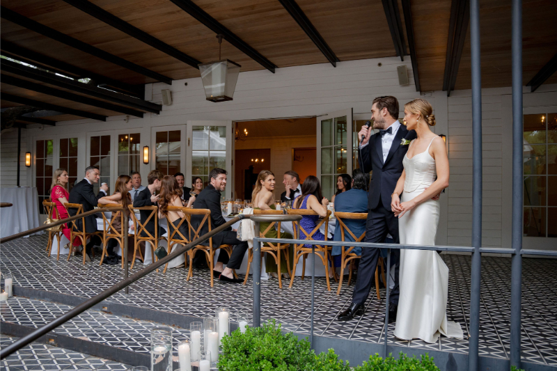 A bride and groom stand together as the groom makes a speech to seated wedding guests at their outdoor reception area at Mattie's Austin.
