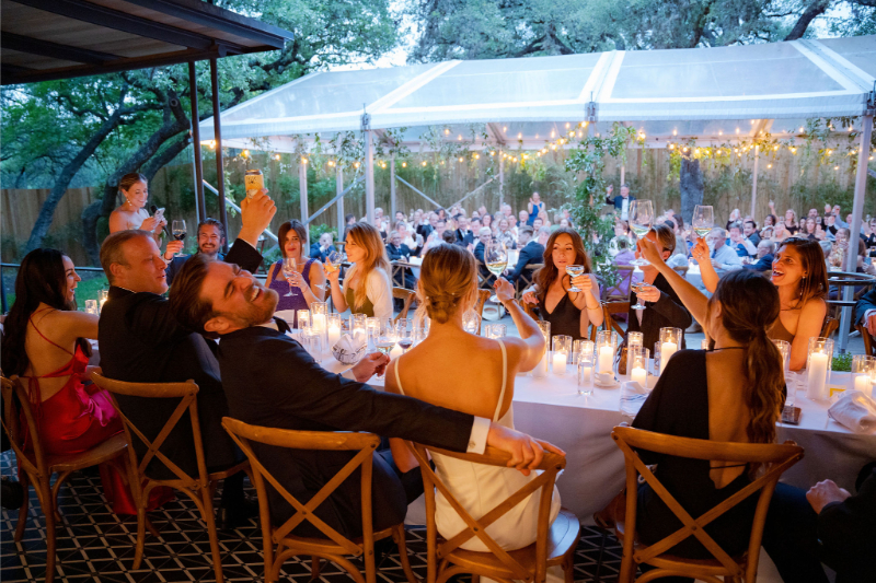 A group of people dressed formally sit at a long table under a tent at Mattie’s Austin, raising glasses in a toast during an outdoor event with string lights overhead.