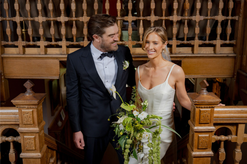 A bride in a white dress holding a bouquet stands next to a groom in a black tuxedo on the wooden staircase at Mattie's Austin, both smiling.