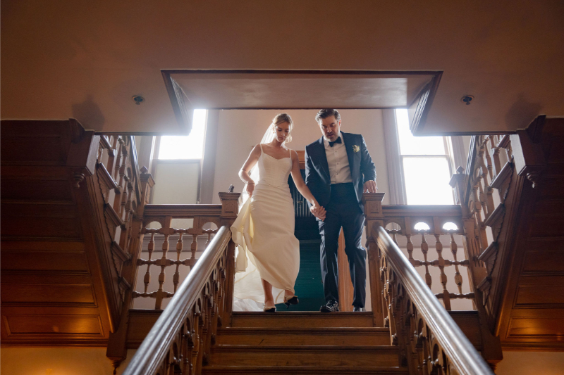 A bride and groom walk down a wooden staircase at Mattie's Austin, holding hands, with natural light streaming through the windows behind them.