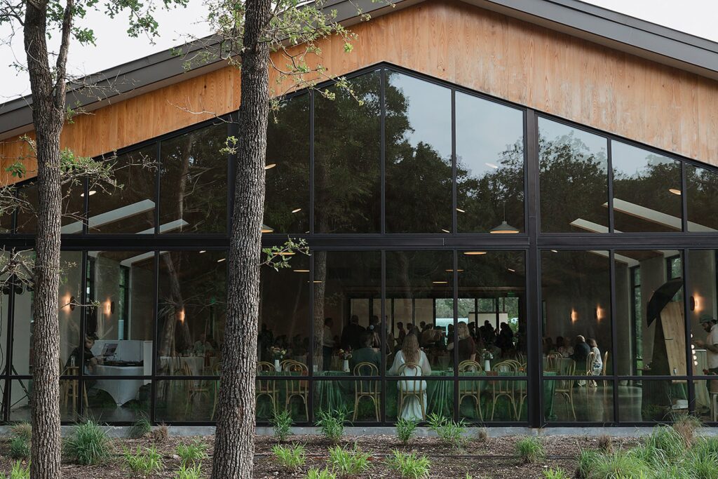 The Grand Lady stands proudly, a rectangular building with large glass windows revealing an event inside. Trees and greenery in the foreground add a touch of elegance to this captivating scene.