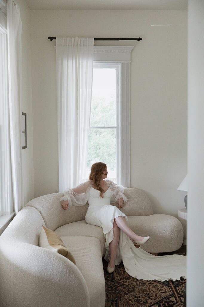 A person in a white dress with sheer sleeves, sits gracefully on a curved, textured sofa. Sunlight pours through large windows onto a patterned rug in the bright room.