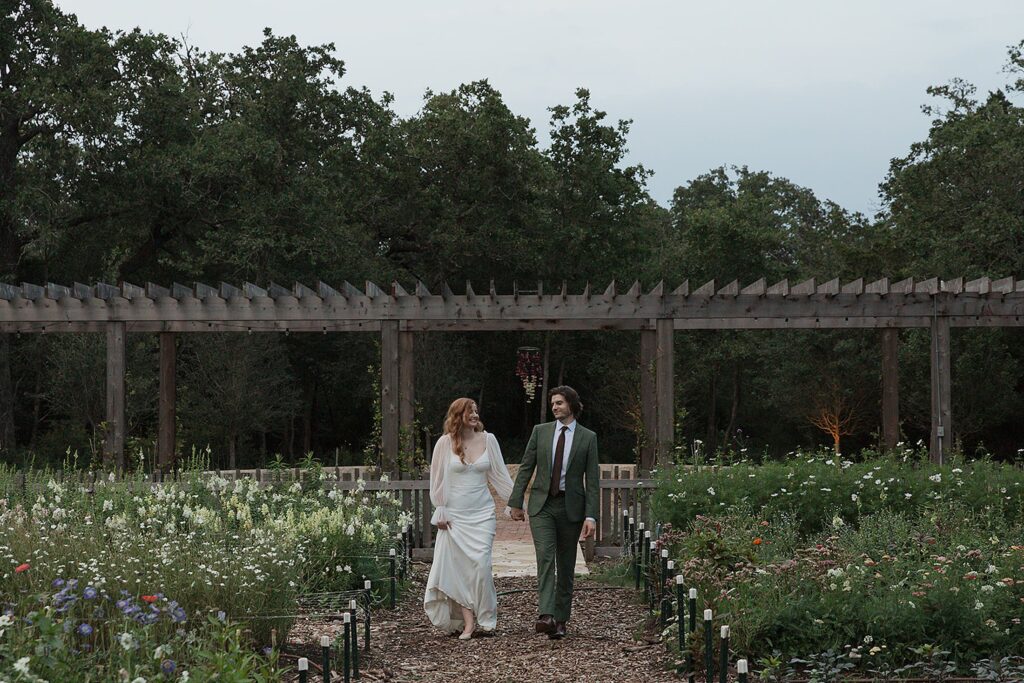 The bride and groom, hand in hand, stroll through the enchanting garden path of The Grand Lady, surrounded by lush greenery and vibrant flowers beneath a charming wooden pergola.