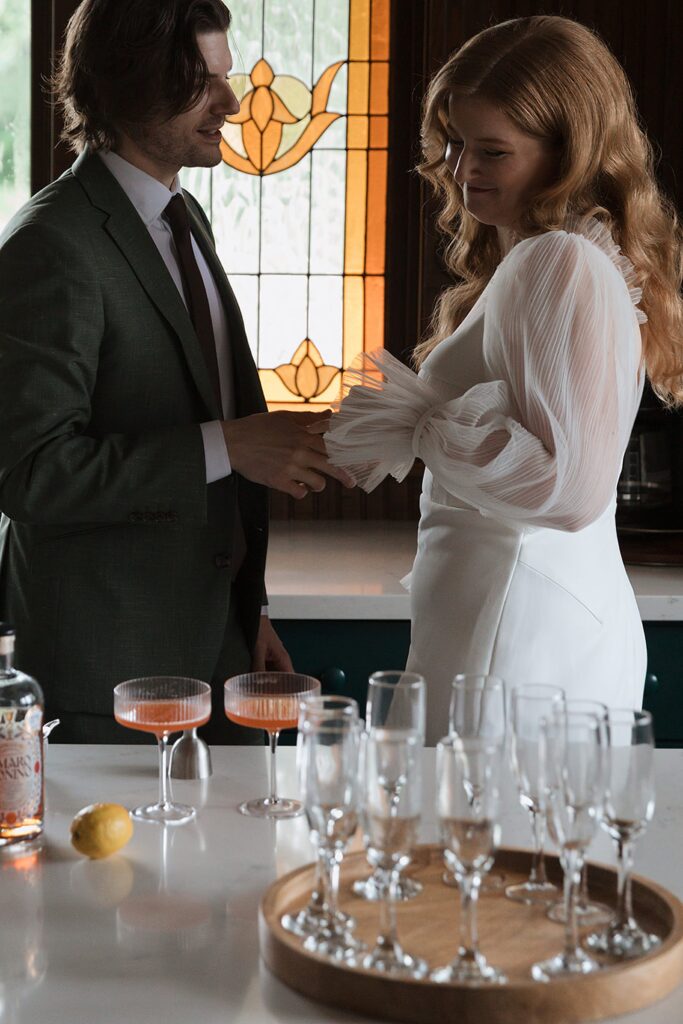 A man and woman stand by a table adorned with cocktails and empty glasses. A lemon and a bottle are also on display, perfectly capturing the essence of The Grand Lady, whose stained glass window adds an air of elegance to the scene.