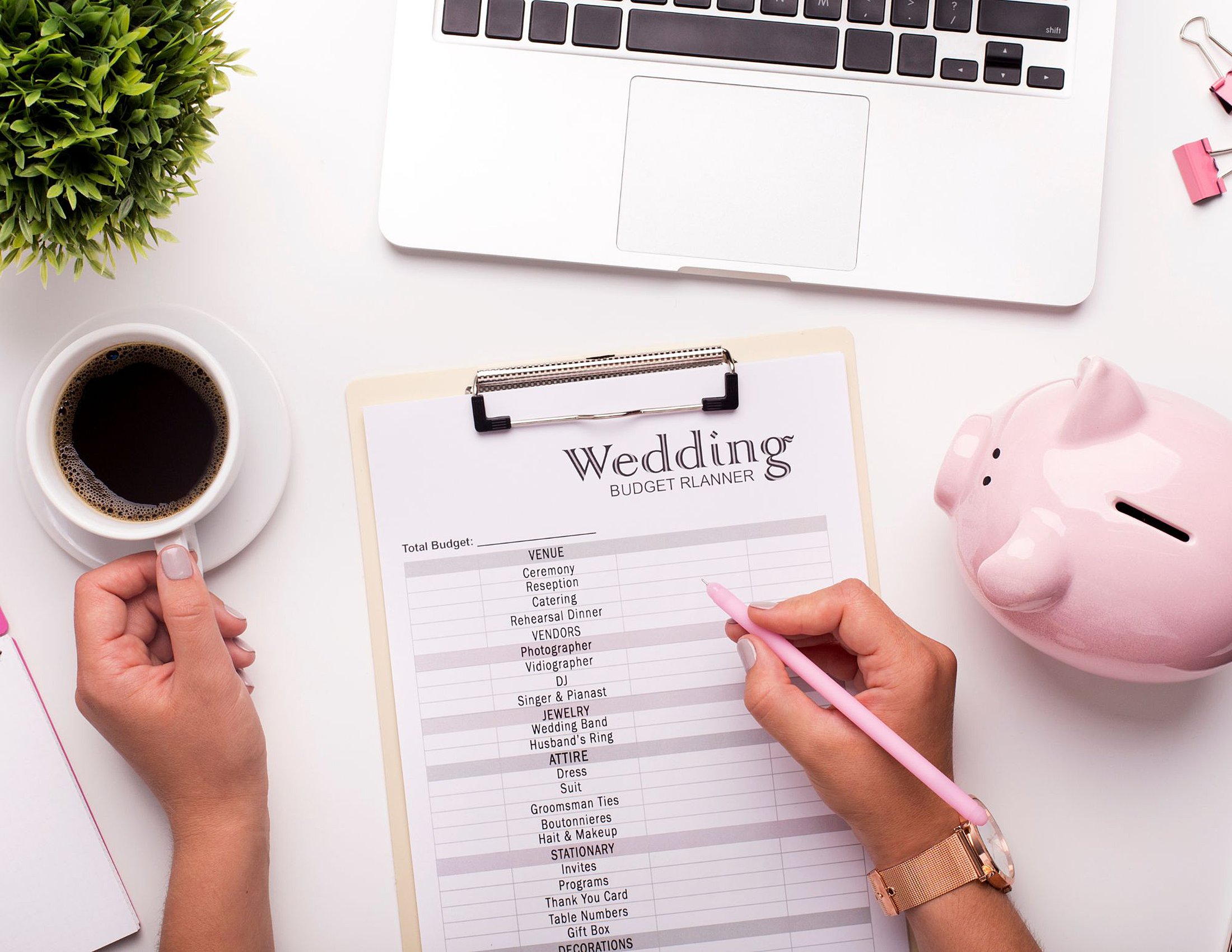 A wedding budget planner on a clipboard sits beside a laptop, a steaming cup of coffee, and a pink piggy bank, all crucial for avoiding common wedding planning mistakes. A person is thoughtfully holding a pen and their coffee, ready to jot down Austin wedding planning tips.