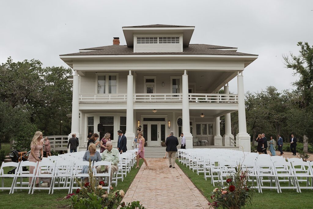 The Grand Lady, a majestic white two-story house, boasts a welcoming porch and rows of white chairs strategically arranged on the lawn. The scene is animated by people mingling, adding warmth and vibrancy to this elegant setting.