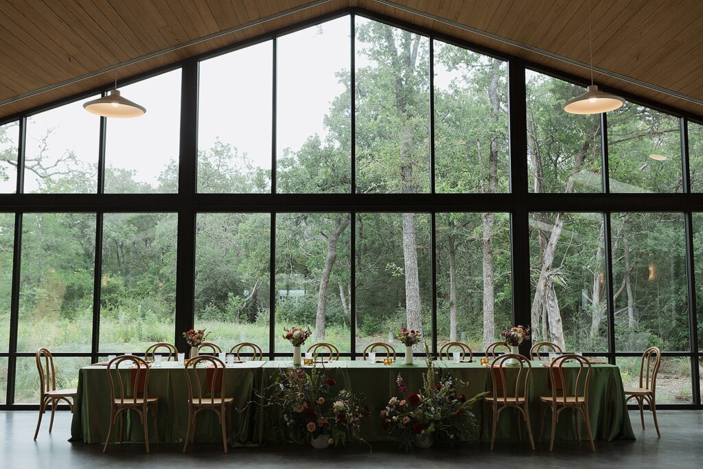 A long dining table, called The Grand Lady, with a green tablecloth is set with chairs and floral arrangements, located in a room with large windows overlooking a wooded area.