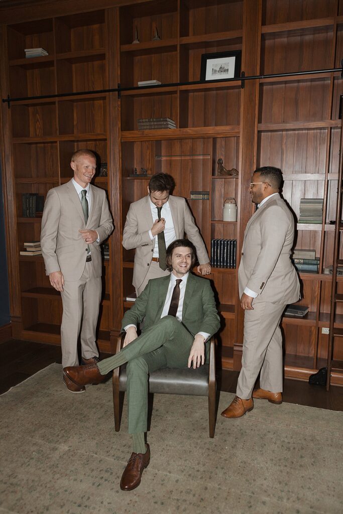 Four men in suits, one seated and three standing, pose in a room with wooden bookshelves.