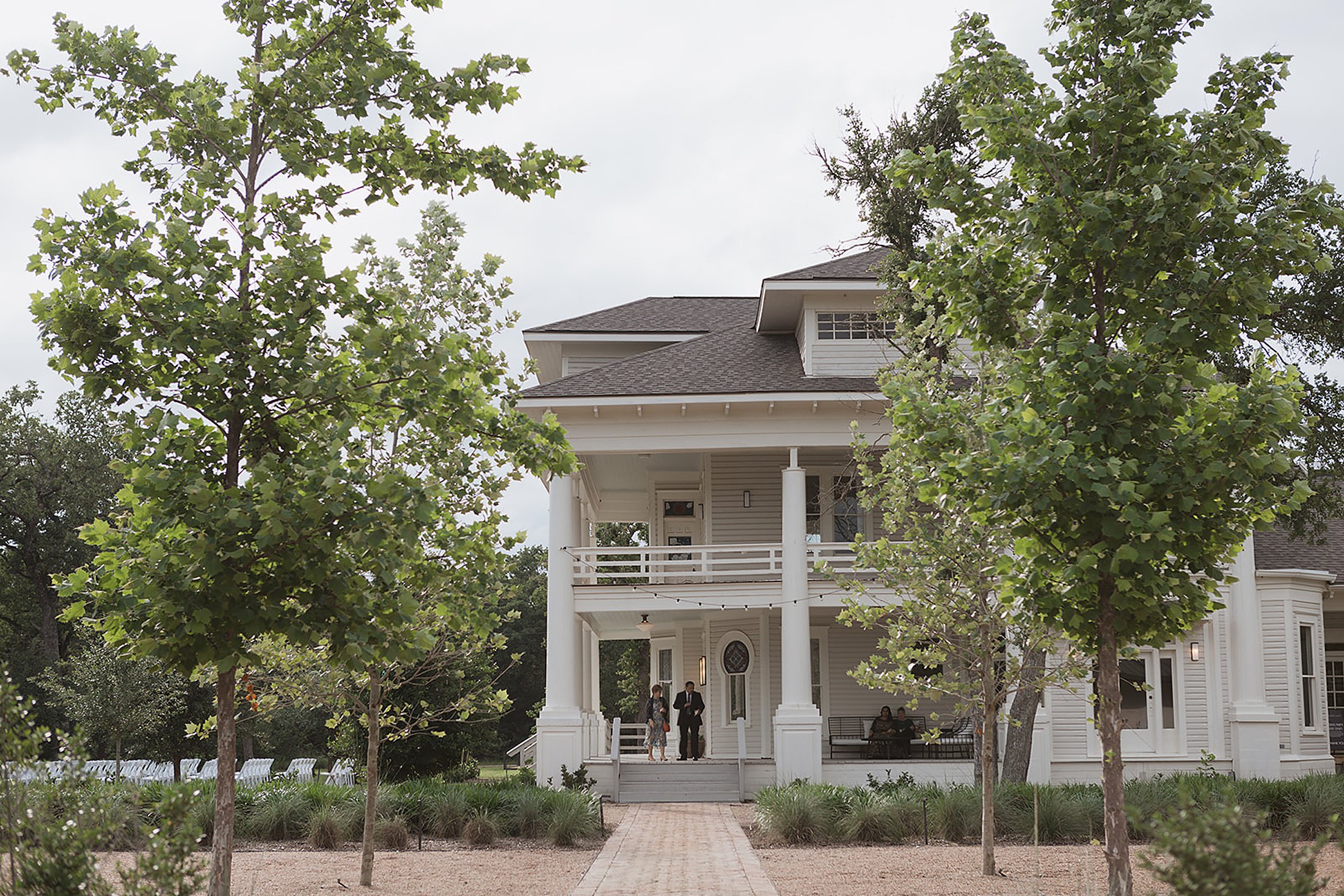 The Grand Lady stands tall as a large white two-story house with a porch, gracefully surrounded by trees and a brick pathway leading to the entrance.