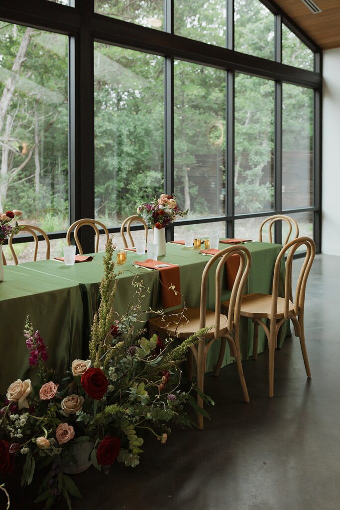 A dining table with green cloth, set with chairs, floral centerpieces, and candles, stands elegantly next to floor-to-ceiling windows. 